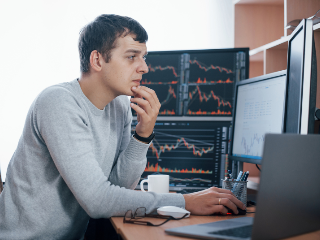 Focused trader analyzing charts on multiple monitors in a home office setting, with financial graphs and stock market data displayed on screens.